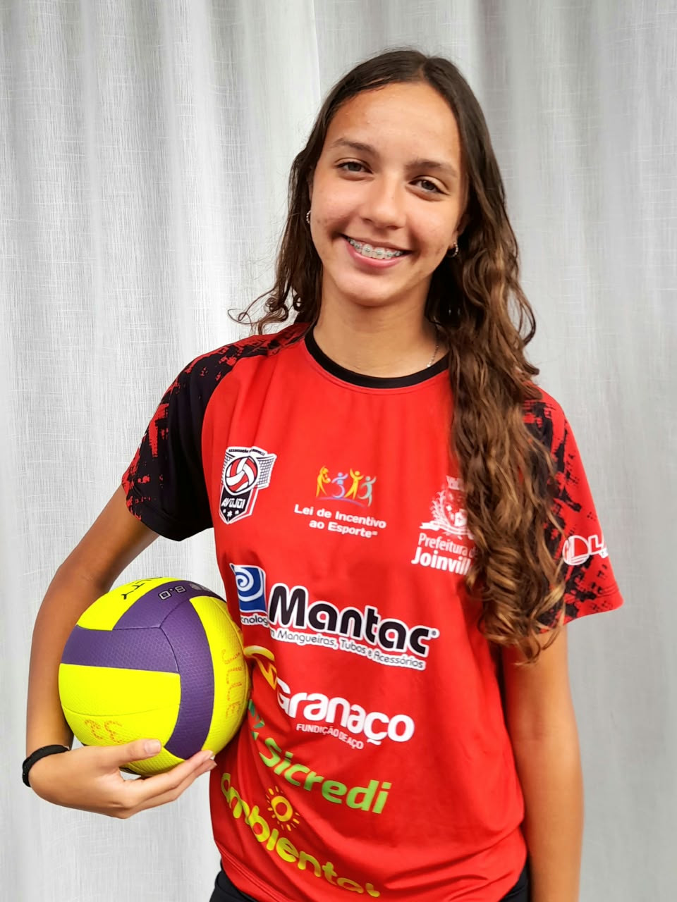 Retrato de atleta com camiseta vermelha de Joinville, segurando uma bola de vôlei.