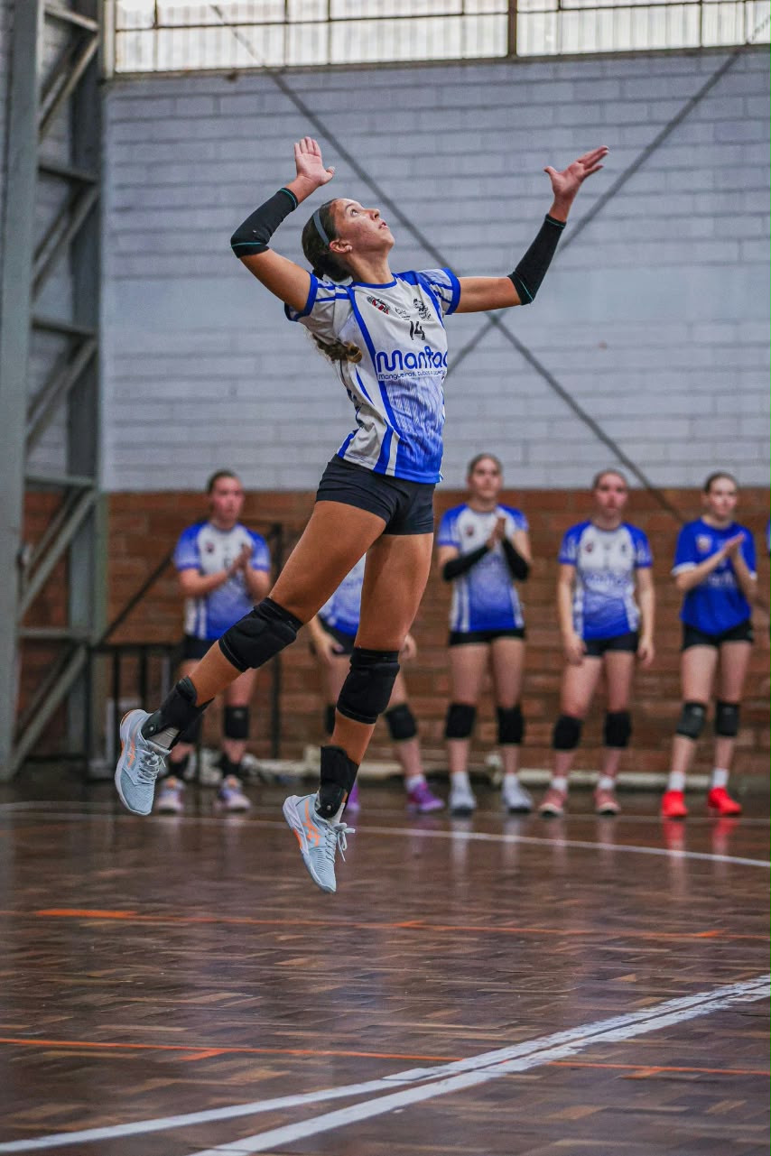 Atleta de uniforme azul e branco número 14 salta em uma quadra de ginásio, com outras atletas ao fundo.