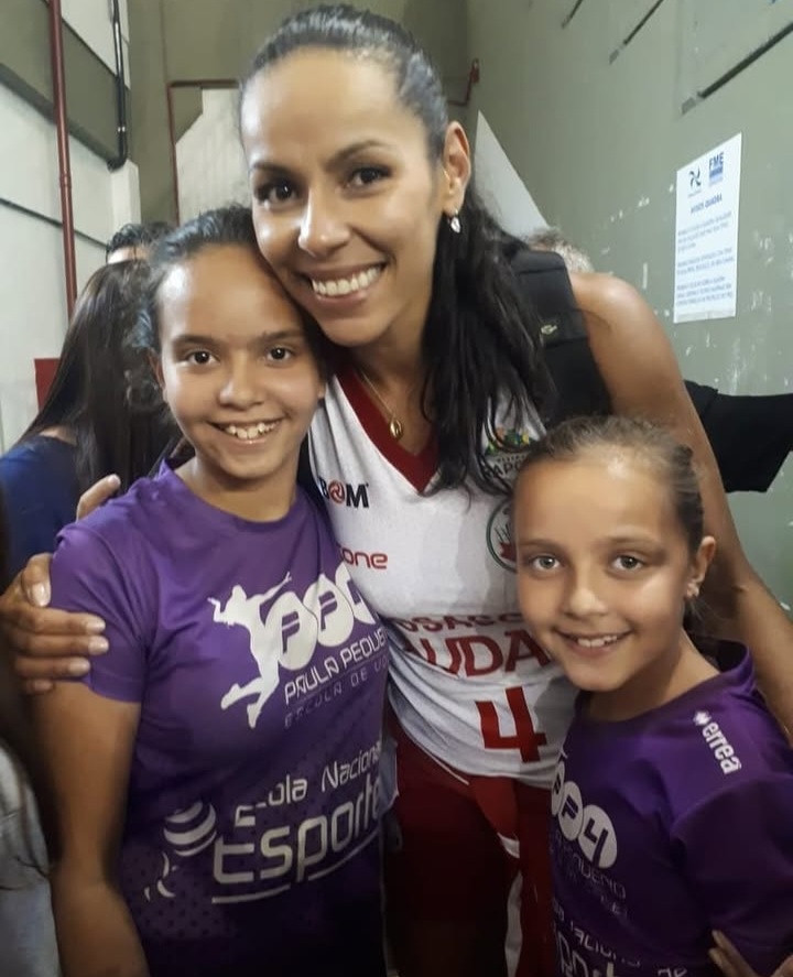 Uma adulta de uniforme esportivo branco e vermelho posa ao lado de duas crianças com camisetas roxas.