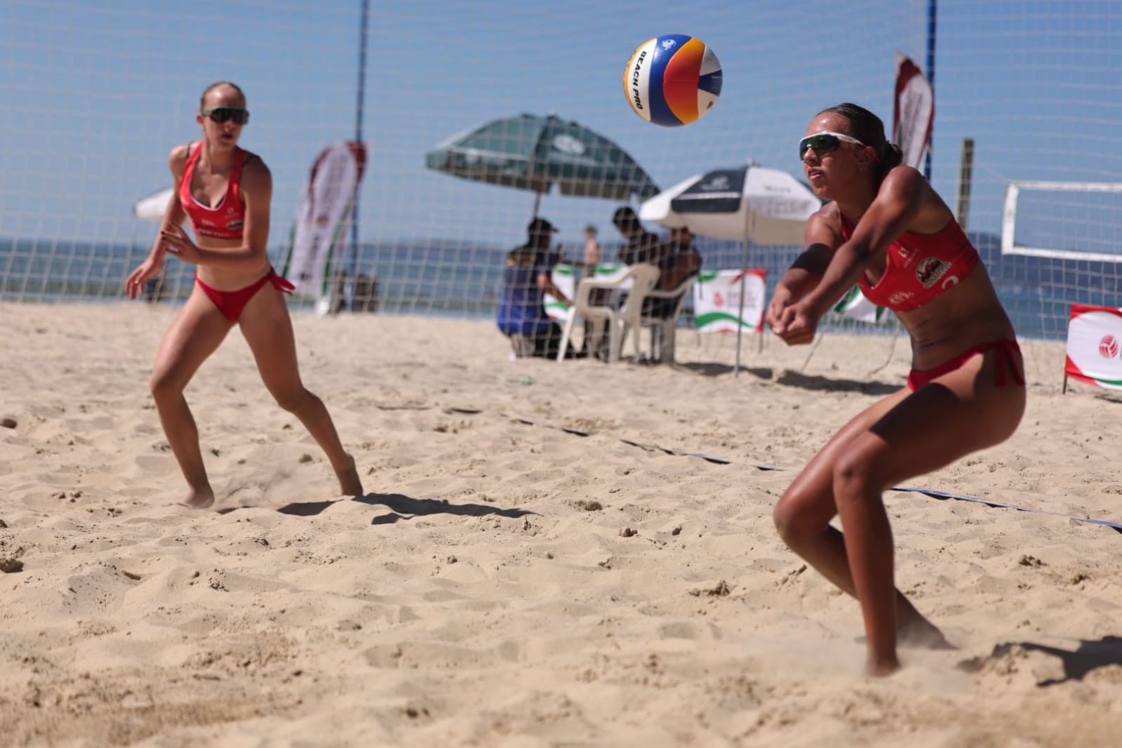 Duas atletas de uniforme vermelho em uma partida de vôlei de praia, com a bola próxima à recepção.