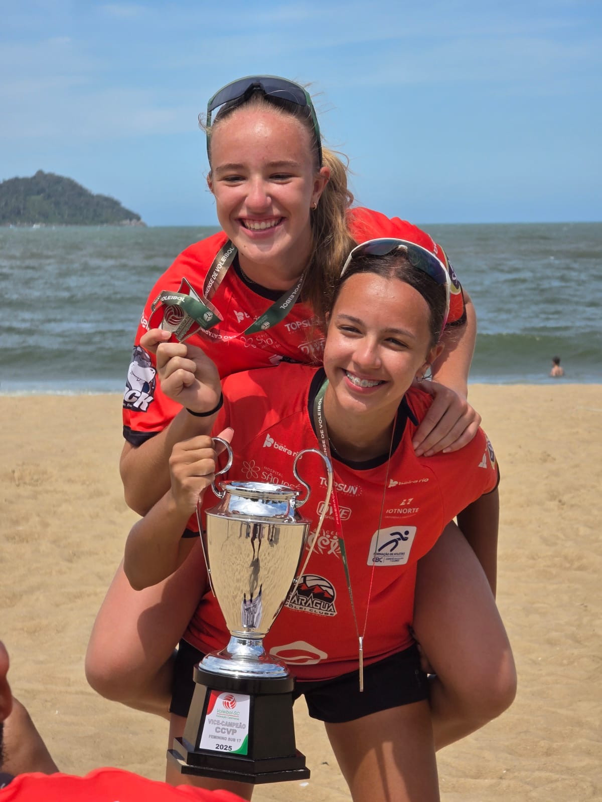 Duas atletas de camiseta vermelha e medalhas comemoram na praia com o troféu de vice-campeão do CCVP feminino sub-17 de 2025.